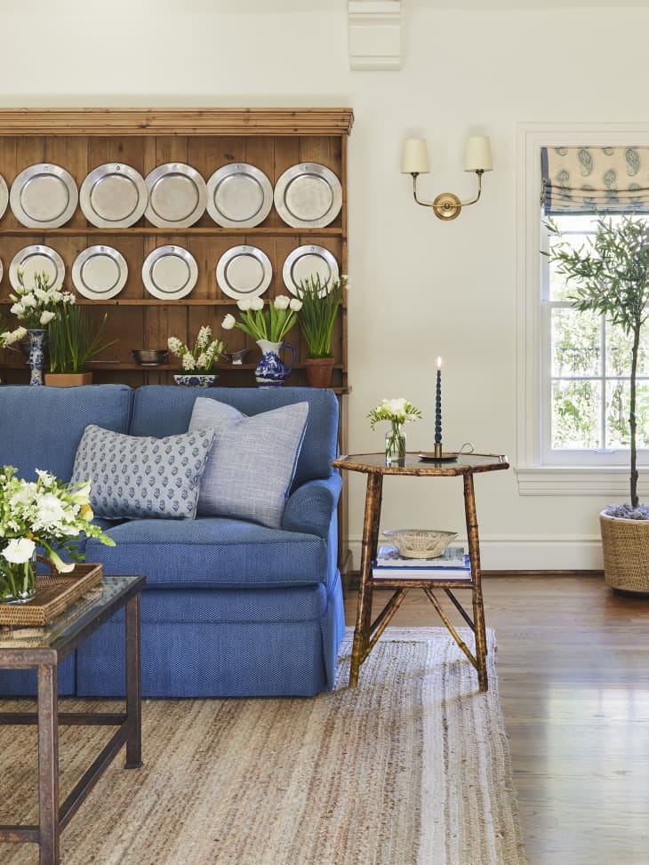 living room with blue sofa, neutral striped area rug, wood wall of shelves with plates, plants, white walls, cream and blue window valances, pale blue and white armchairs