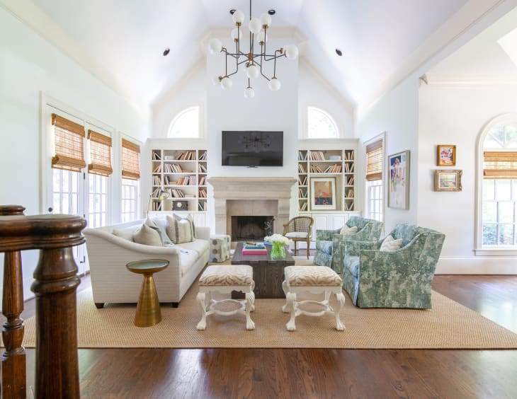 Living room with pale neutral sofa, wood block coffee table, 2 teal and cream floral armchairs, 2 wood (painted white) legged ottomans, pale stone/brick fireplace, white walls, angled arched ceiling detail, modern chandelier, 2 tall bookshelves flanking fireplace