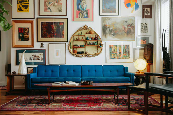 A blue mid-century sofa on a burgundy rug in front of a gallery wall