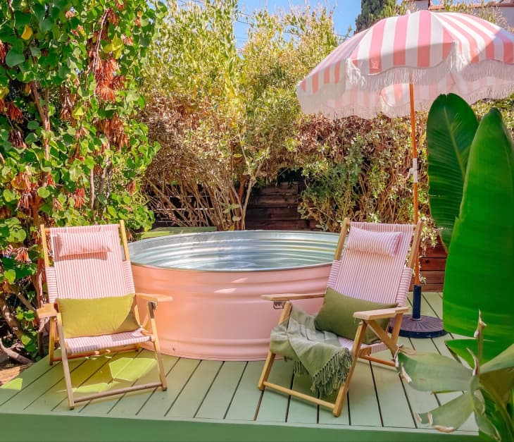 Dani Dazey designed patio area with pink stock tank pool, pink and white striped fabric and wood beach chairs, pink and white umbrella with white fringe, trees and plants around