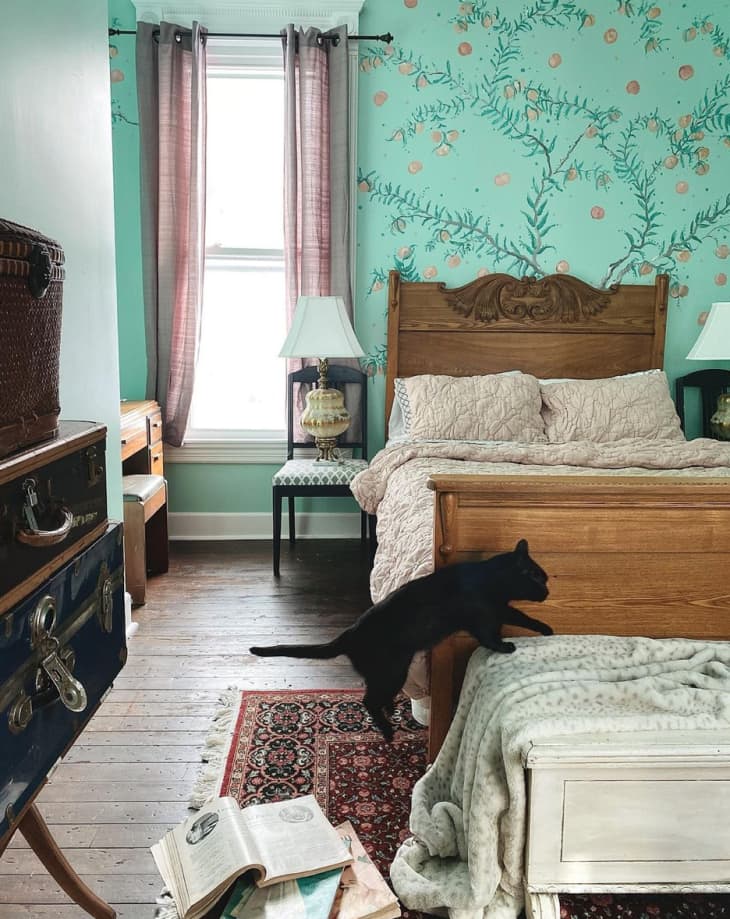a mint green patterned wallpaper with a wooden antique bed. a black cat jumps on a chest in the foreground
