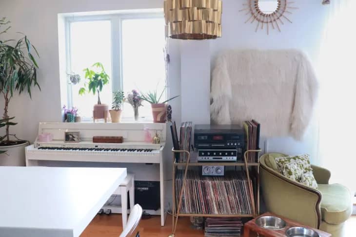 an apartment wall with a turntable, records, a white piano and a TV in the background with a blanket draped over it