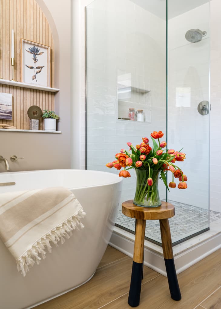 wood floor, soaking tub, standing shower, arched hanging shelves, wood stool with bouquet, white tile, glass shower walls and door