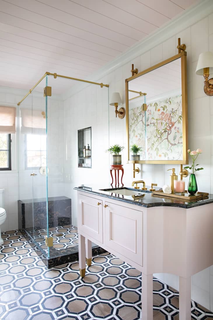 hexagon pattern tile, brown white and black hexagon pattern tile floor, pink vintage inspired vanity, dark counter top, glass doors on standing shower, black ledge at bottom of shower, paneled ceiling, gold fixtures, large gold square mirror, shaded sconces, built in shower seat, shower cubby