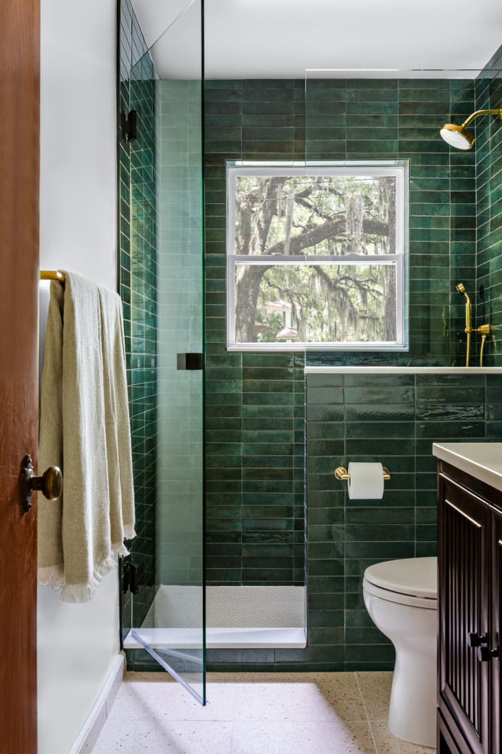 dark green subway tile on shower wall to ceiling, pony wall next to toilet, glass shower door, walk in shower, gold fixtures, towel bar, dark vanity, white countertop