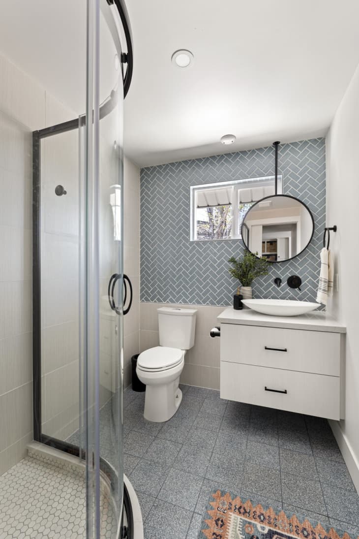 light blue grey square tile floor, curved glass wall of standing shower, white penny tile in shower, blue penny tile backsplash behind sink and toilet, white floating vanity, round mirror, sink bowl, plant, coral multi colored bath mat