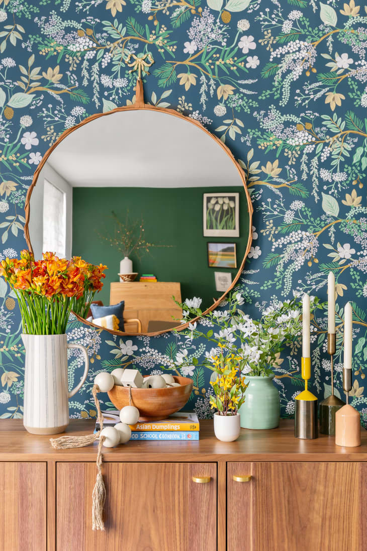 detail of large wood credenza with large white pitcher of orange flowers, stack of books, wood bowl, other plants and candles, round wood frame mirror on blue floral wallpaper wall,