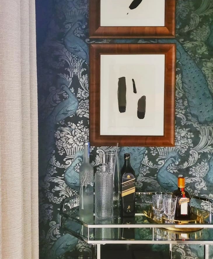 Bar cart with bottles, gold tray, glasses, wall behind has blue floral and bird print wallpaper, framed art