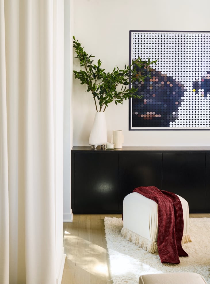 Room with off white walls, wood floor, black credenza, small cream ottoman or stool covered with fringe-trimmed fabric, shag rug, plant in white vase