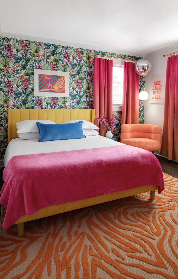 Bedroom with orange textured tiger stripe rug, bed with yellow fabric headboard, White duvet with hot pink throw, bright blur throw pillow, orange cushioned accent chair, sunset gradient curtains, green and red botanical wallpaper