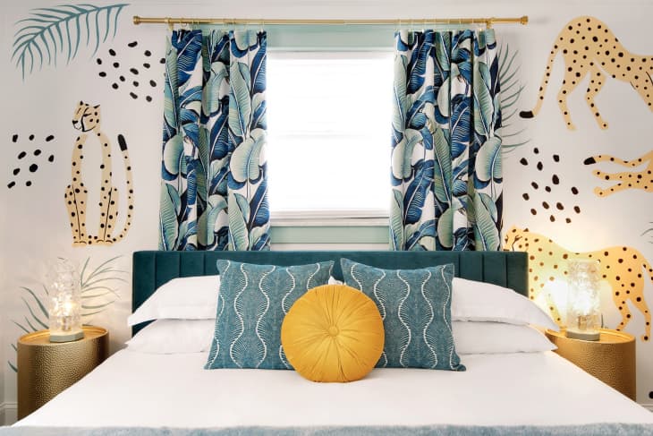 Bedroom with cheetah wallpaper, tropical leaf teal and white curtains, bed with teal quilted headboard, white bedding, teal and gold pillows, 2 gold bedside tables with glass lamps