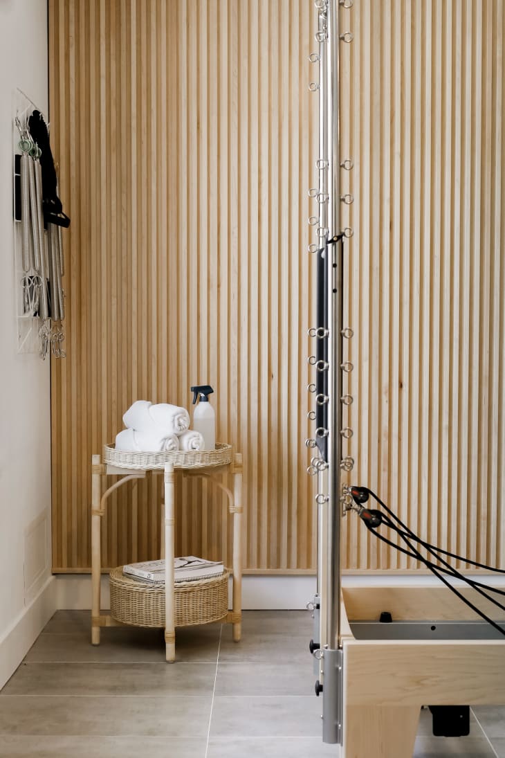 corner of room with wood and wicker tiered table with rolled spa towels, spray bottle. Decorative wood slat wall behind