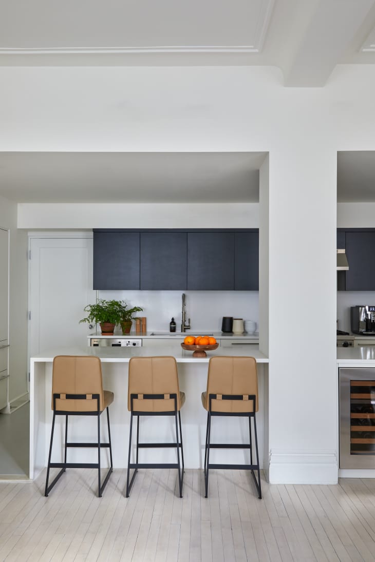White kitchen with navy blue cabinets, large white kitchen island with 3 tan leather stools with black legs, painted slat wood floor