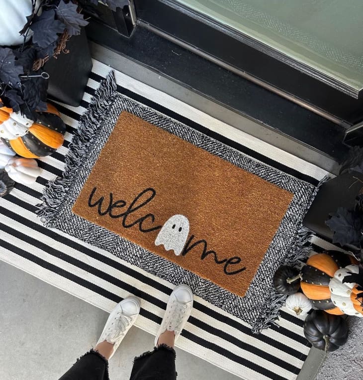 Front door welcome mat with a ghost on it, surrounded by other pumpkin/halloween decor