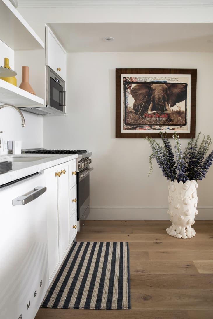 wood floors, black and white striped rug, white cabinets, large floor vase with plant, elephant art, microwave, oven, sink, open shelving