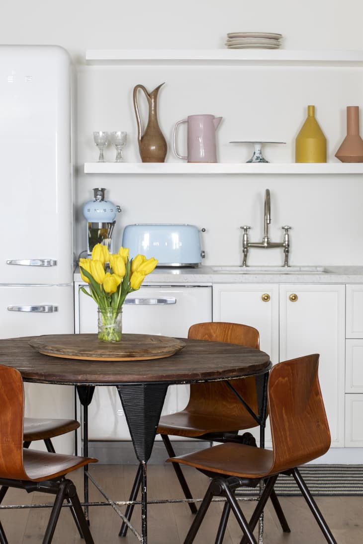 round dark wood table, dark orange curved chairs, white cabinets, yellow pink and orange pottery jugs, open shelving, vintage white fridge, sink, baby blue toaster, baby blue smeg machine, yellow flowers