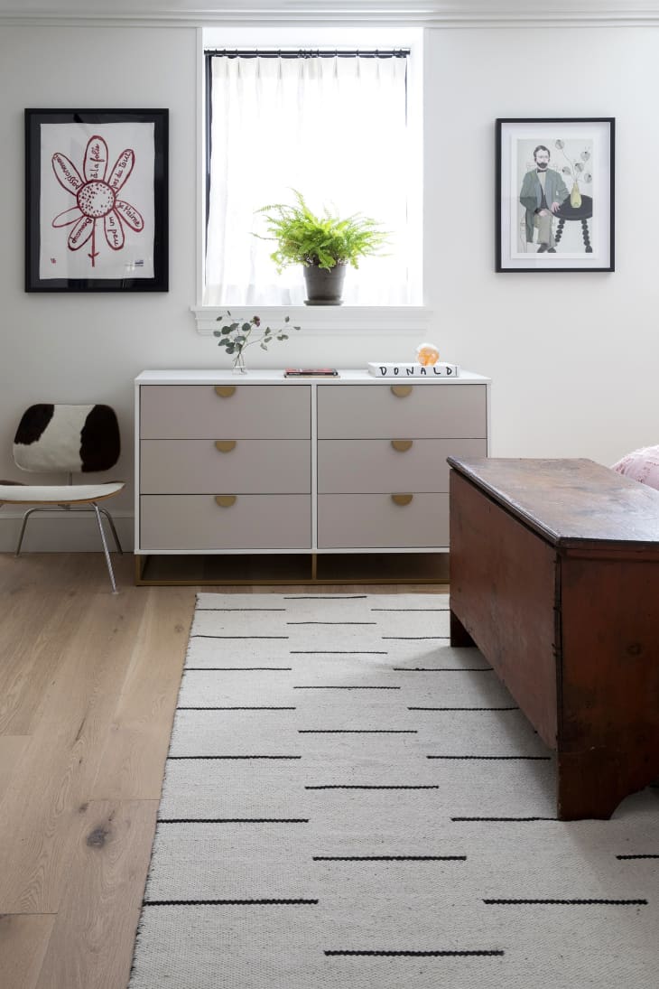 wood floors, whtie dresser, cow print desk chair, wood chest, beige rug with small black dash mark lines, plant in windowsill, art