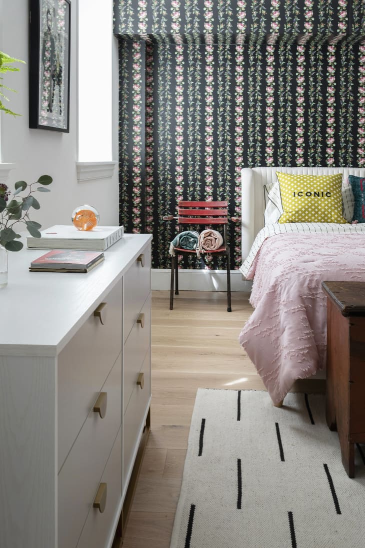 wood floors, wallpaper striped pattern accent wall bleeding up on to ceiling, white dresser, pink throw, yellow pillow, white and black thin line pattern rug, red patio chair