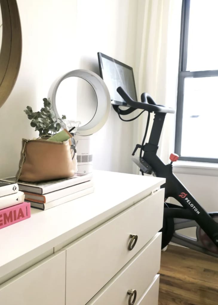 peleton bike in the corner of room next to white dresser and window