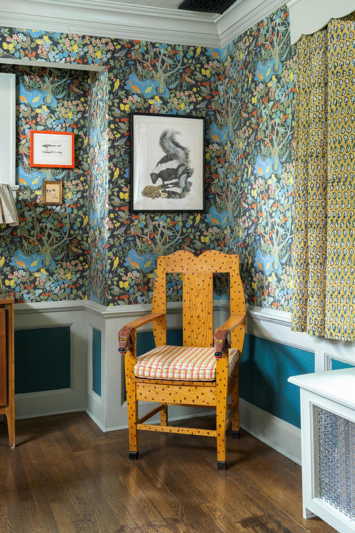Wooden chair in corner of room with patterned wallpaper and short curtains on window.