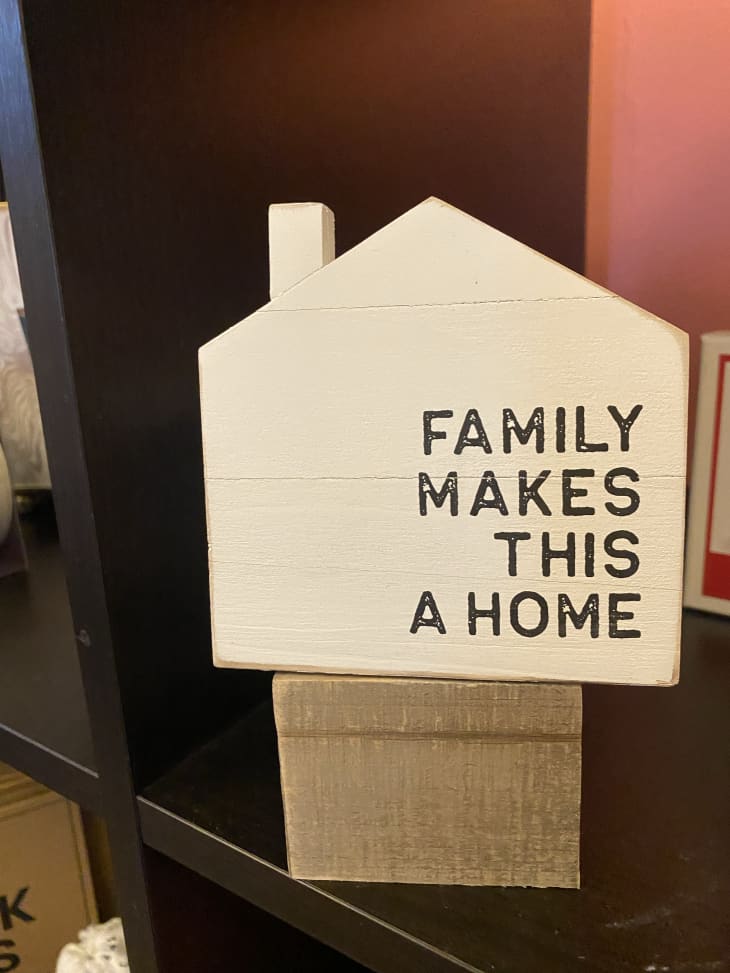 wooden sign in the shape of a house with words stenciled on that say "FAMILY MAKES THIS A HOME"
