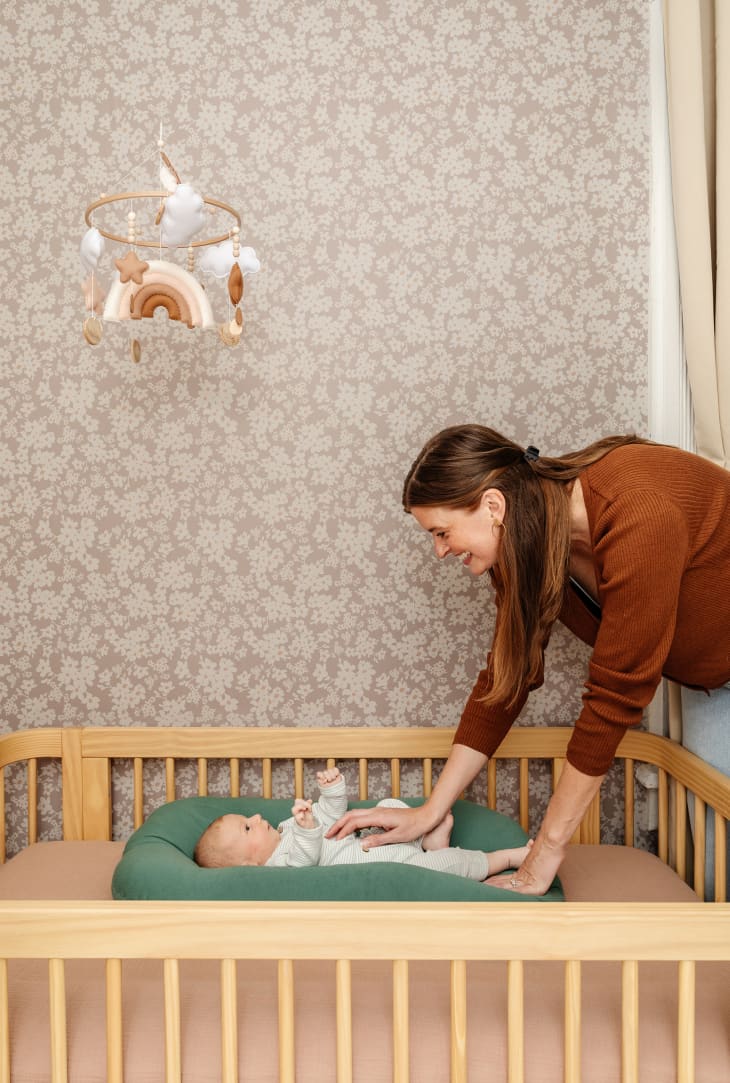 Mother bent over crib to comfort baby in newly decorated nursery with botanical wallpaper.