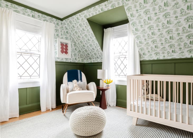 Nursery with green and white forest wallpaper and green wainscoting with white curtains and cozy chair