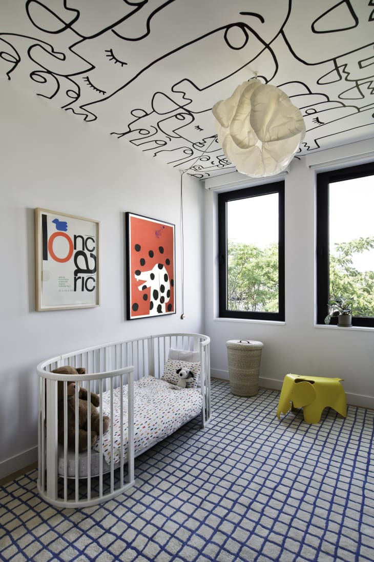 black doodle line wallpaper on ceiling, oval white crib, black and white checkered carpet, yellow stool, bold art, paper chandelier