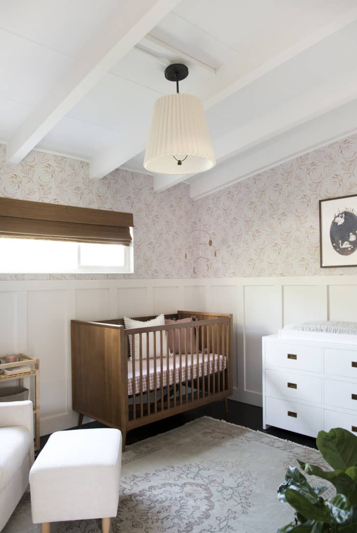wood crib, half wall wainscoting, half wall pink floral wallpaper, white dresser and ottoman, bamboo roman shades