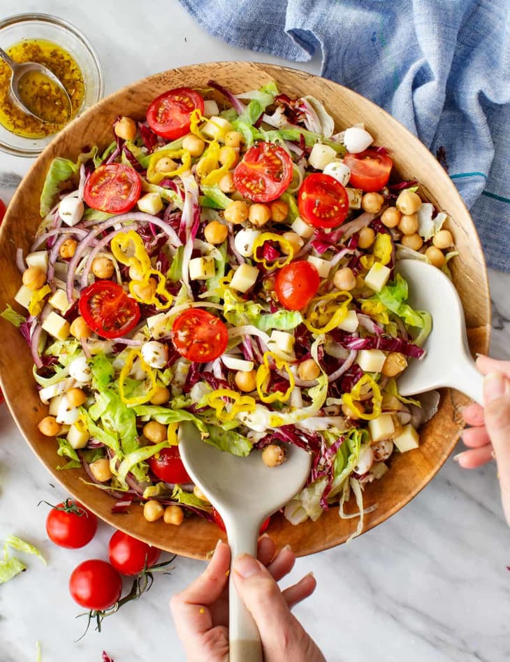 chopped salad with tomatoes, chickpeas, and veggies in a wooden bowl. two hands holding white tongs.