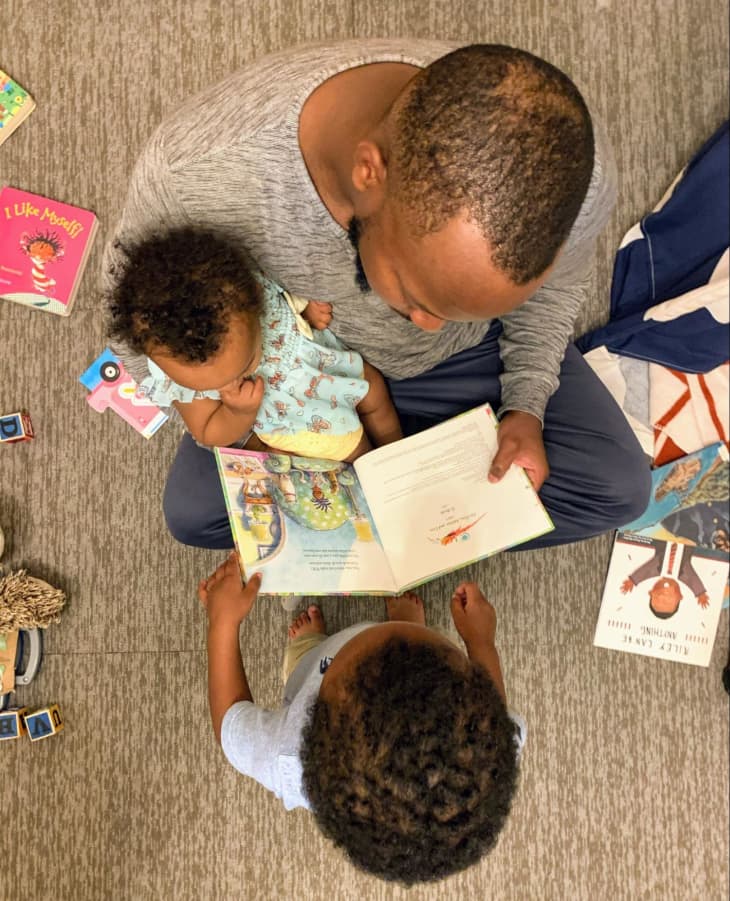 Father reading to baby and young child. Photographed from above with picture books scattered throughout the image.