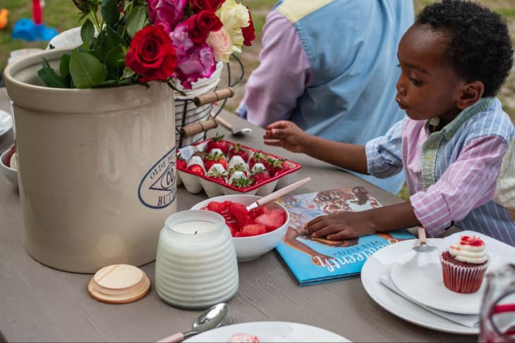 Young toddler boy in a pink and white striped shirt and blue vest is sitting at a table outdoors, reaching for a skewer of strawberries. In front of him: a white plate with a single cupcake, candles, and a big flower pot filled iwth pink and white flowers. An adult sits in the background, in clothes matching the young child's.