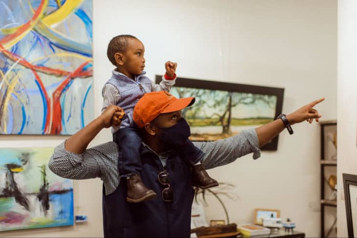 Father and son at an art gallery. Toddler son is on father's shoulders ad father points to a piece of art.