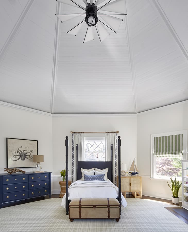 vertical shot of a nautical themed bedroom with a geometric fan resembling sails of a ship. four post bed in center of frame with trunk at the foot of the bed. on the righthand side, a wide window with a striped valence. on the lefthand side, a navy nightstand.