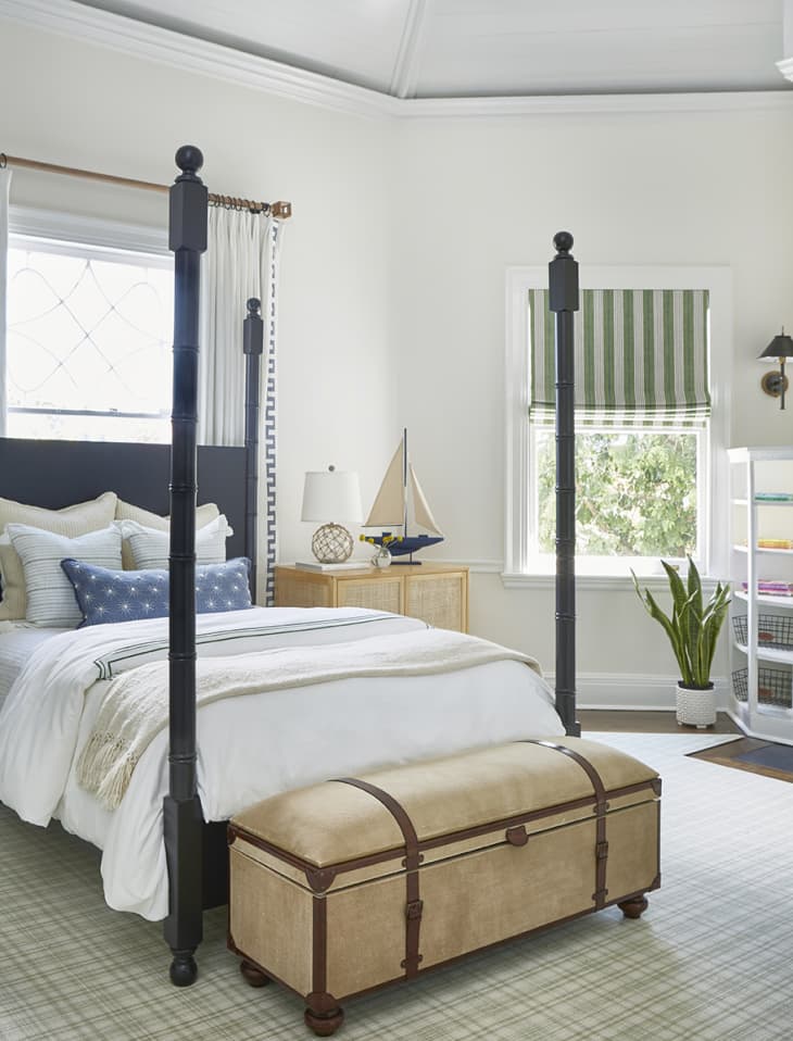 nautical themed bedroom with four-post bed, made up with white sheets and blue and white accent pillows. trunk at the foot of the bed. bright light coming through windows, as well as a striped valence.