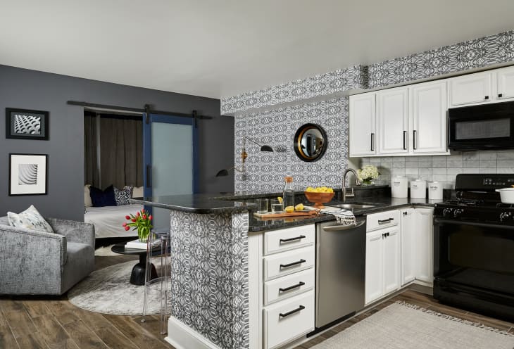 Modern looking kitchen overlooking a living area. Black and silver appliances, white cabinets with silver fixtures, and black and white modern wallpaper.