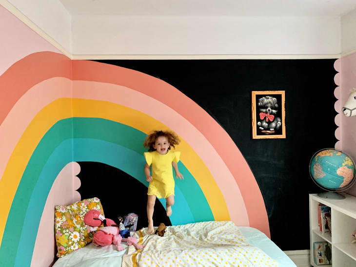 Bright rainbow painted in corner of a wall in the bedroom, spanning two walls. Five stripes of teal, green, yellow, dark pink, and light pink. Little girl's bed in center, with girl jumping on bed in a yellow dress and a big smile.