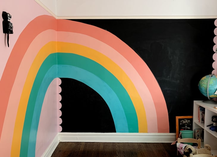 Bright rainbow painted in corner of a wall in the bedroom, spanning two walls. Five stripes of teal, green, yellow, dark pink, and light pink. Walls behind the rainbow are black and pink, with pink scallop detail.