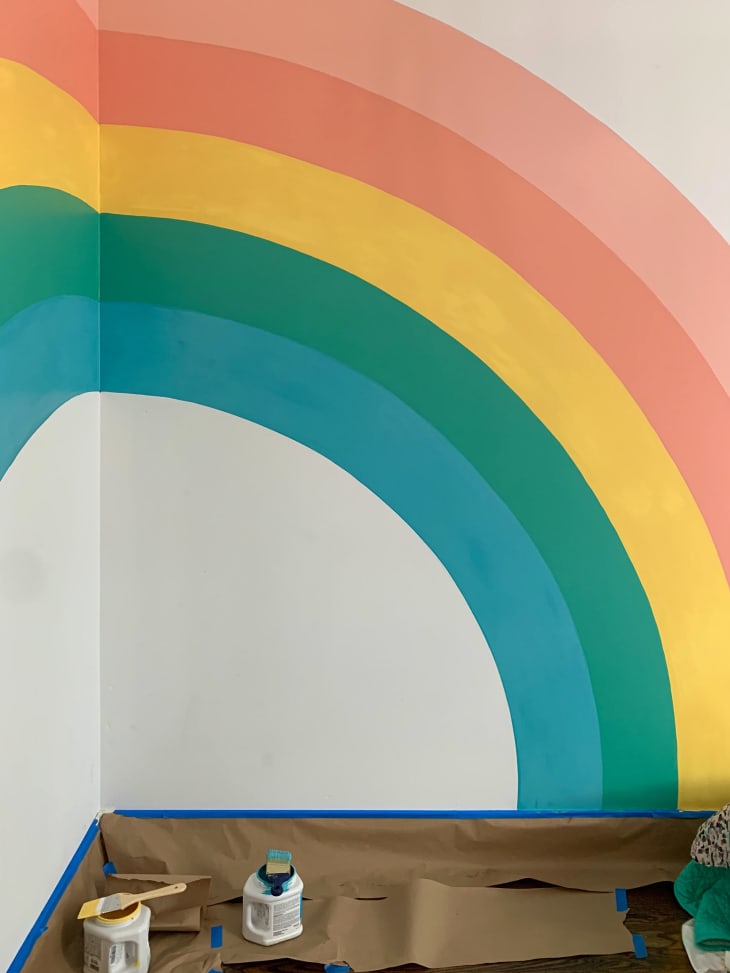 Bright rainbow painted in corner of a wall in the bedroom, spanning two walls. Five stripes of teal, green, yellow, dark pink, and light pink. Wall behind the rainbow is white.