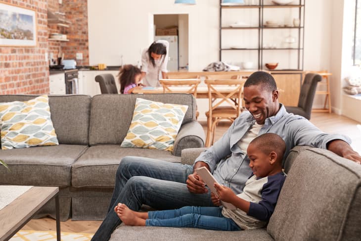 Black father and son sit on couch with an iPad. Both are smiling. In the background, a woman leans over a young girl to work on homework at a kitchen table. The scene is bright and happy.