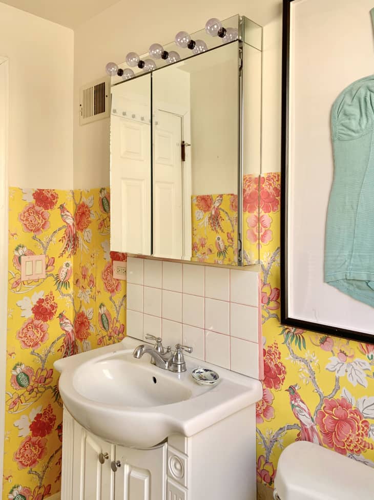 beautiful canary print wallpaper covering half the wall. top half is white paint. Backsplash with custom pink tile behind the sink. Above the toilet, a large framed turquoise vintage bathing suit.