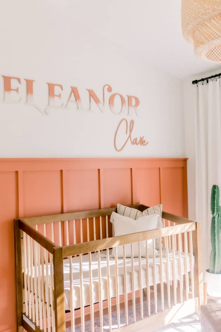 Neutral wall with coral wainscoting, with crib in front. Name on crib in coral ombre reads "Eleanor."