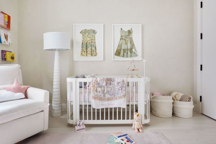 Light neutral nursery with large open windows. White crib sits below two framed pictures of dresses on the wall. On the right, two cloth baskets for toys. On the left side of the frame, a white glider under a bookshelf.