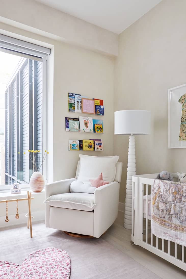 Light neutral nursery with large open windows. Glider angled near window below books. Large table lamp next to crib with a quilt hanging over the side.