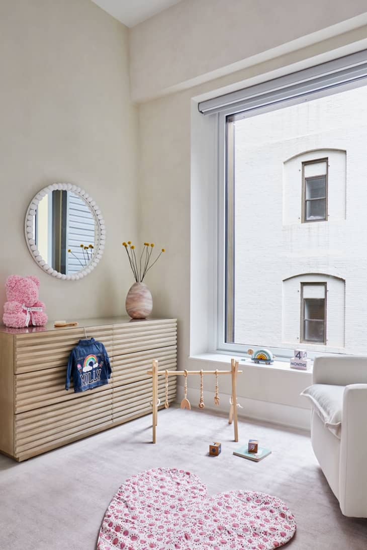 Light neutral nursery with large open windows. Light pine dresser with circular mirror on the wall. On the floor, a heart-shaped play mat for a baby, as well as a small baby play set.