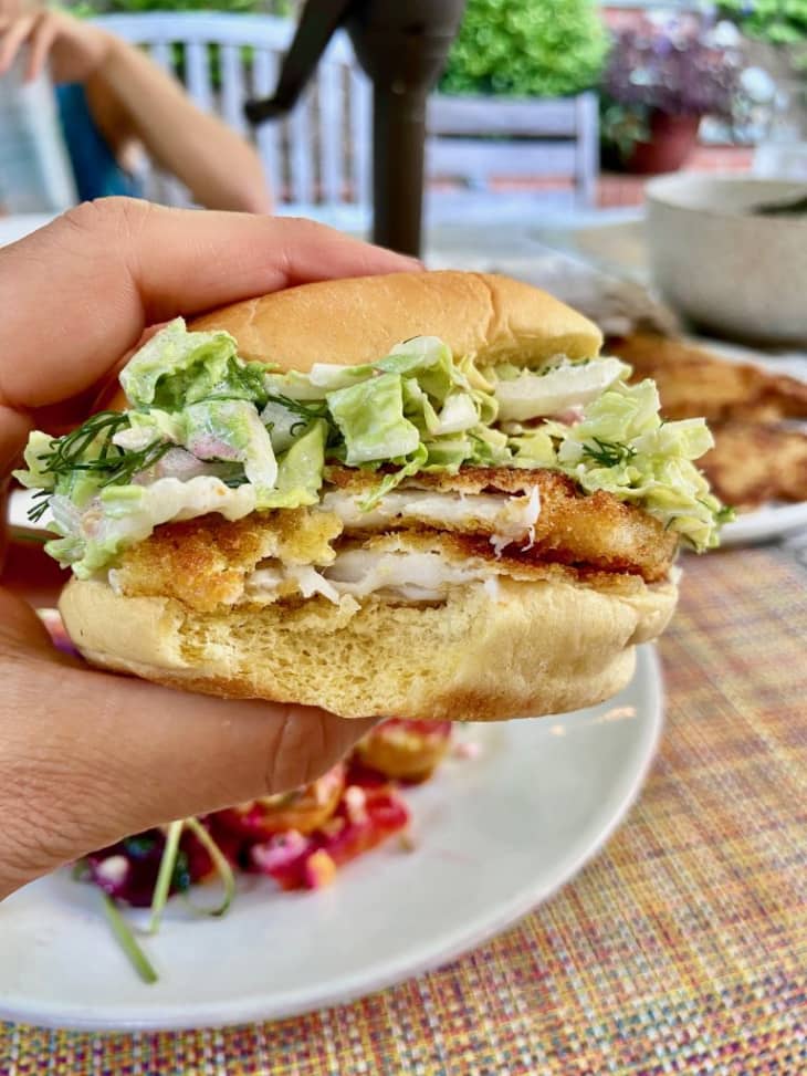 view of a filet of fish sandwich that someone is holding in their hand. On bite has been taken. other plates and people at table. outdoors