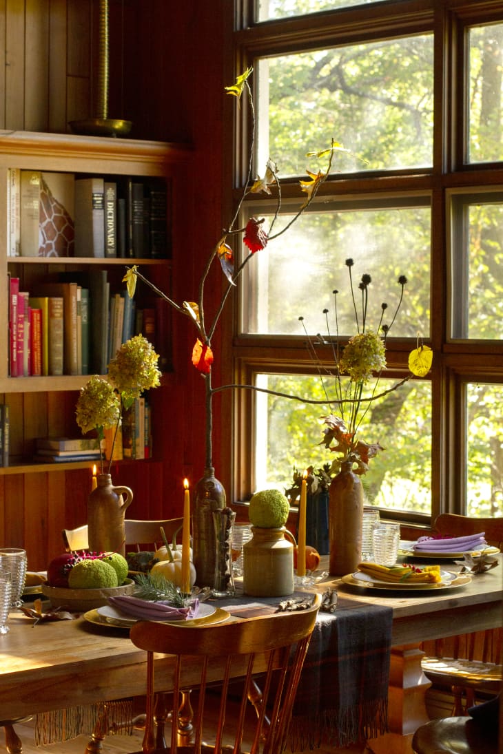 Thanksgiving table next to a large window. Table has a lot of decorative elements--candles, a vase with a branch that has leaves with names on them, a couple of floral arrangements
