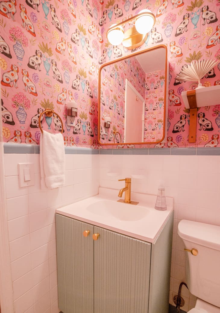 bathroom sink with light green cabinet. Gold hardware, including faucet, cabinet pulls, towel ring on wall. Mirror with gold frame, and wall sconce with gold details. Pink wallpaper illustrated with dogs. White toilet with gold flush handle
