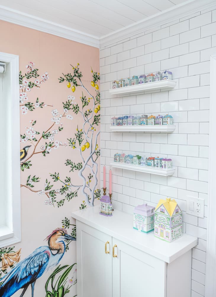 corner in kitchen with pink wallpaper with an illustration of a heron and flowers/leaves. White cabinet with shelves above, decorated with small ceramic houses
