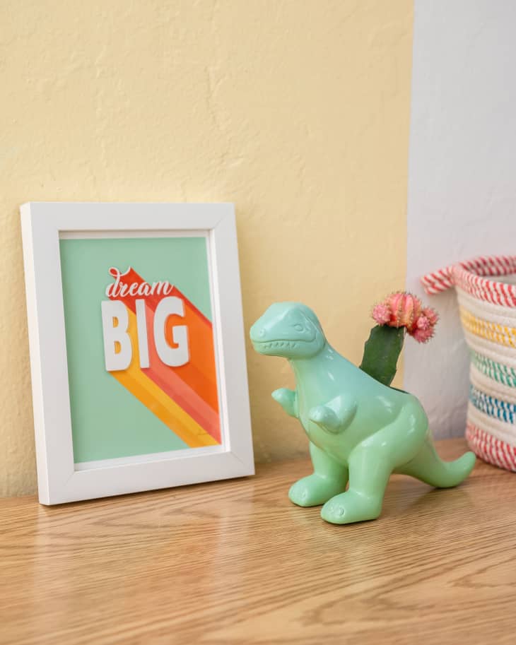 shelf with framed print that says "dream big". Green dinosaur figurine next to it, and striped basket partially in frame